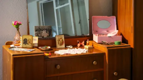 Museum of the Home A room set in 1956 which has a dressing table on display that has items including a small vase with a pink flower, old photos, a pair of glasses and a pink make up case