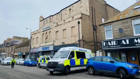 BBC Police at scene of search warrant being executed in Peterhead's Queen Street, blue and yellow checked police vehicle in street outside shops and buildings.