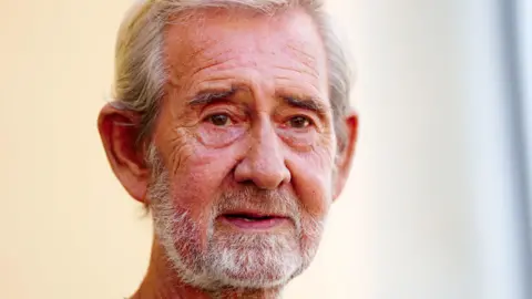 David Hunter looking to the right of the camera. He has white, short hair and a white beard. He has a slightly furrowed brow and his mouth is partially open.