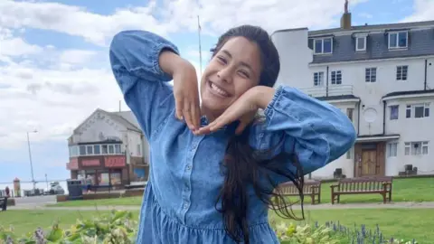 Handout A teenage girl with long glack hair, posing with her hands up to her face. She is outside and wears a long-sleeved denim dress.