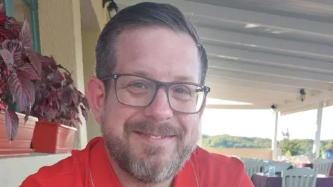 Essex Police Andrew Rayner is in just off centre to the left looking towards the camera smiling. He has short dark grey hair and is wearing glasses and a red T-shirt. In the background are rows of tables and chairs.