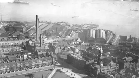 A black and white aerial view of Hamilton Square surrounded by Georgian houses and a town hall. The roofs of the rail station platforms can be seen in the background by the River Mersey where a few vessels are sailing.