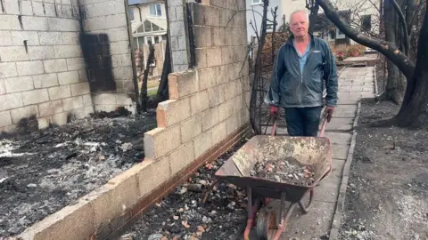 A person stands beside a wheelbarrow filled with rubble next to the fire-damaged remains of a building. Charred walls, burnt trees and ash-covered ground surround the site, while neighbouring homes can be seen in the background.