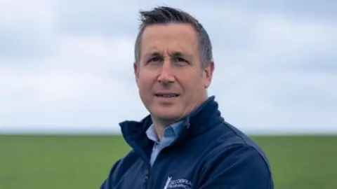 Tom Barker A man stands in a green agricultural field and looks slightly over his shoulder to look at the camera. He is slightly smiling. He has short dark hair with some grey in it. He wears a navy blue jumper with a lighter blue shirt underneath. 