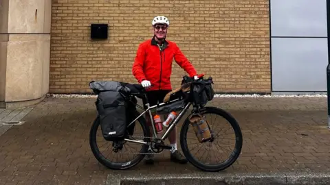 Norman Brown A man with a bike stands outside a brick building. He is wearing a red top and black shorts and has a helmet and sunglasses on.