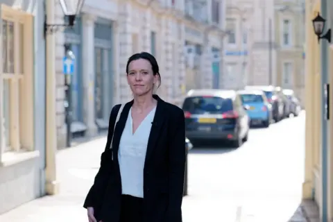 Laurence Cawley/BBC Lucy Davies walking towards court. She has dark brown hair tied back in a ponytail. She is smartly dressed with a white blouse and a dark suit jacket with a bag over her right shoulder.