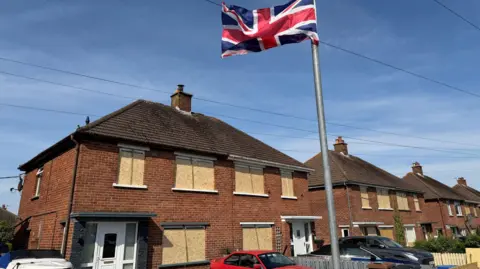 a street of semi-detached houses with cars parked outside and a Union flag flying on a lamppost. Most of the glass on the houses (with a few exceptions, like glass next to front doors) has been covered with wood.