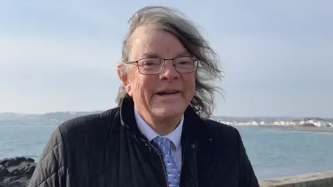 Man in his late 50s, early 60s with thin grey and semi-long hair stood in a shirt and tie. He is wearing glasses and behind him you can see St Peter Port. 