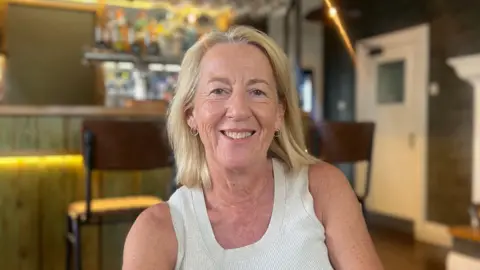 Blonde lady wearing a light gray vest smiling at the camera. She is sat in front of a pub bar, and there are bar stalls on both sides of her.