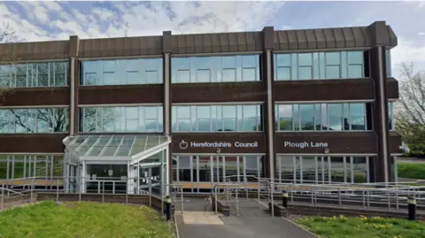 The front of Herefordshire Council's building. It is brown with several large glass windows. There are metal handrails outside the front doors and green grass
