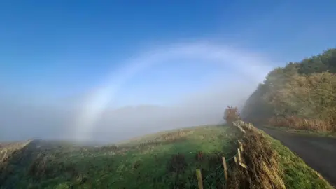 Kim Bennett A grey circular arch of cloud over a path and a hillside. There is green grass on either side. The sky above is blue.