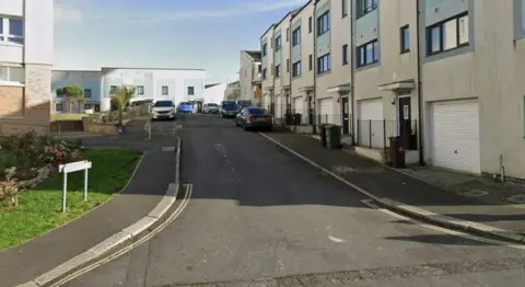 A street view of Curtis Street in Plymouth. There is a row of houses, with garages at the bottom on the right, and houses at the end of the road. On the left is a street sign on grass and a building on the left. 