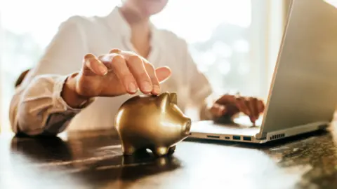 A woman's right hand touching a small golden piggy bank in the foreground. Her left hand is on a laptop keyboard in the background.