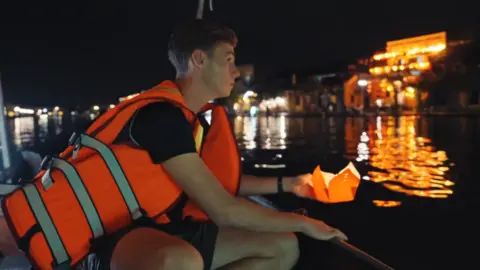 Studio Lambert Alfie Watts, wearing a life jacket, leans out of a boat as he prepares to place a lantern with a candle in it on the water