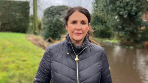 Tracie Cadman, a woman with brown hair, is wearing a black coat and is standing outdoors with a flooded road behind her.