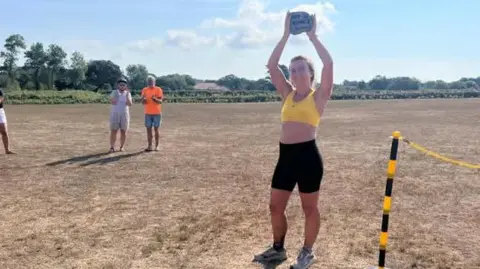 Emily Torode Emily Torode wears a yellow top and black leggings as she holds the trophy above her head, while two people are seen clapping in the background 