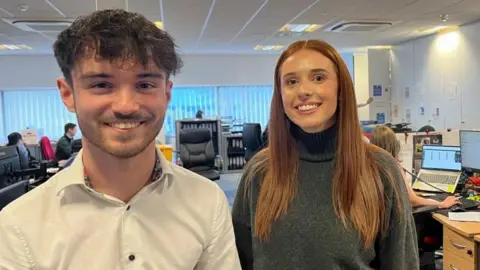 Andrew Turner/BBC Toby Lockyer is wearing a white shirt which has black buttons. He is smiling, has a close cropped beard and long dark hair. Next to him is India Peak, who has long auburn hair, and is wearing a grey polo-neck jersey. They are both standing in the office of Stephenson Smart, where they both work as apprentices in accountancy.