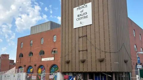 A large red brick building with multiple sections set back from one another and a ramp to the right side. There is a sign above the glass entrance doors which reads Prince of Wales Centre. The section to the left of the entrance has a number of archways containing colourful murals.