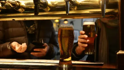 PA Media Customers collecting their drinks in a pub from the bar where you can see the golden pumps which pour the beer.