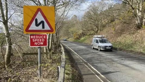 A country road with a van passing and a reduce speed sign