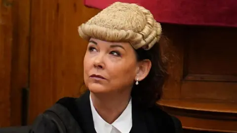 The Lord Advocate,  Dorothy Bain KC, in formal legal attire sits in a courtroom, wearing a traditional barrister’s wig and pearl earrings. She looks slightly upward to her left with a composed, thoughtful expression. Behind her are wooden panels and a red fabric backdrop.
