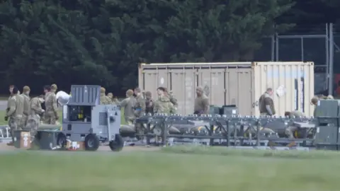 EPA Image of an air base, with a shipping container in the background. In the foreground can be seen a number of military personnel in khaki uniforms who appear to be checking explosive ordnance.
