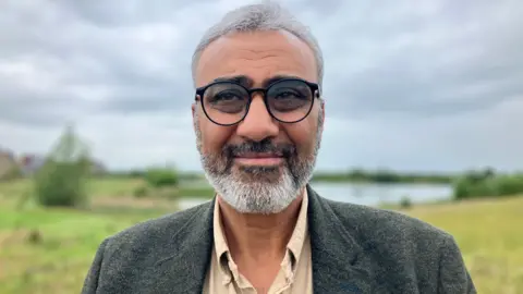 JOANNA TAYLOR/BBC Jawad Nawash smiling and looking just past the camera. He is wearing a beige shirt, dark green blazer and glasses. Behind him is a lake surrounded by grass, out of focus. 