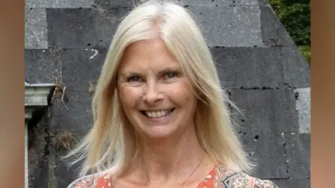 Joy Beresford Frye a lady who is standing outside in an orange dress. She has blonde hair and is looking directly at the camera and smiling.