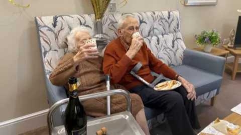 Hambleton Grange Care Home Tony and Joy sit side-by-side on a grey sofa, patterned with purple flowers. Tony is an older man with short white hair, and glasses. He wears a dark orange jumper. Joy is an older woman, with short white hair. She wears a brown knitted jumper dress. They both hold a plastic cups, and Tony has a plate of cake on his knee. In the foreground, an open bottle of prosecco can be seen. 