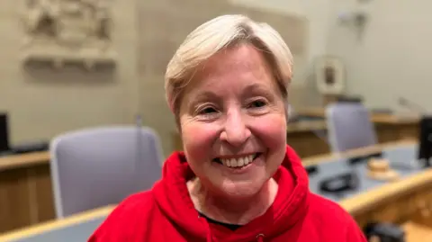 Valerie has short blonde hair and is smiling at the camera. She is wearing a red hoodie standing in a council focus. The benches and chairs behind her are out of focus.
