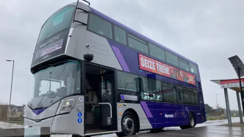 Andy Trigg/BBC A purple and grey electric double-decker bus parked at a bus stop on a rainy day. The bus door is open and a promotional sign on the side of the bus reads 'seize them'.