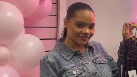 A woman with long black hair is standing between a pink balloon arch and a rack of sparkly jeans. She is wearing a denim jacket top and is wearing her hair slicked back.