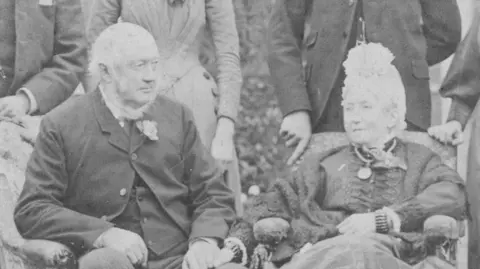 A black and white image of farmer John White (left) and his wife Sophie White (right). They are dressed in Victorian clothing. 