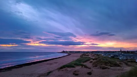 Loki's Keeper Sunrise over houses next to a broad section of dunes leading onto a sandy beach and sea shore. The light is dim with blue sky shading into pink, and purple, with bright gaps where the low sun shines through.