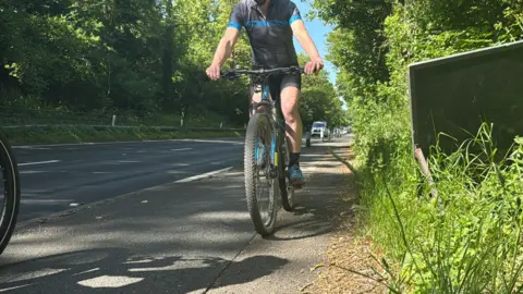 A cyclist on a path rides alongside the traffic on the two-lane A27 near Falmer. To the right of the path are vegetation and a temporary highway sign. 