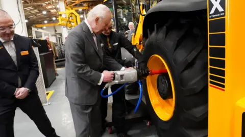 PA Media King Charles wearing a grey suit stands next to two other men and next to a large tyre fitted to a large yellow vehicle. He is holding a piece of equipment as he fits the tyre to the vehicle.