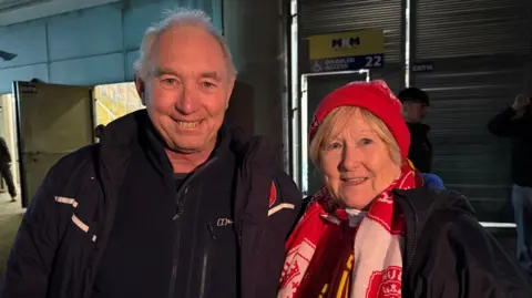 A man and woman stand together smiling. The woman wears a Hull KR supporters hat and scarf, the man has short grey hair and wears dark coloured outdoor clothing.