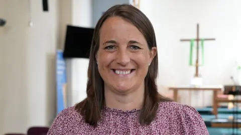 Shaun Whitmore/BBC A woman with shoulder-length hair smiles at the camera. She has brown hair and brown eyes and is wearing a burgundy and white patterned top. Behind her is a simple altar with a wooden table and a cross displayed on a white wall. 