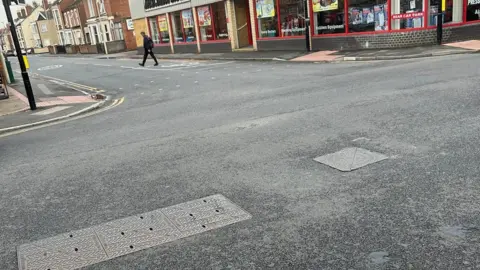 LDRS Clos-up of the manhole on a road near shops and man walking across street.