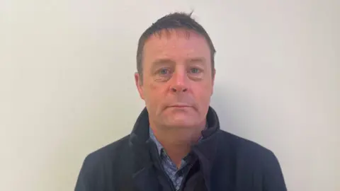 Raymond Gormley looking at camera, wearing a navy coast and a light blue shirt with diamond pattern. 

He is standing in front of a white wall. 