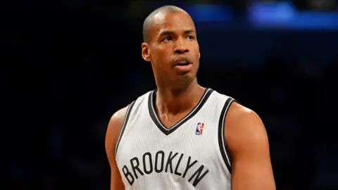 Jason Collins is seen playing for the Brooklyn Nets in 2014. He is wearing a white NBA jersey that reads 'Brooklyn' and is exhaling while looking away from camera as he walks down the court against a black bokeh.