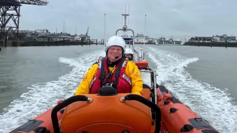 RNLI Andy Gissing wears a helmet and lifejacket at the helm of an orange RNLI rib in a harbour.
