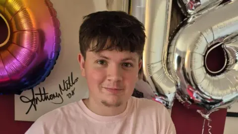 A teenage boy with short dark hair smiles at the camera while wearing a pink t-shirt and standing next to balloons in the shape of the number 18.
