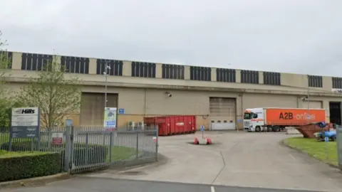 Google Earth A beige, industrial building with a grey sky in the background. There is a red metal container unit and an orange and white HGV. In front of the building is an open metal fence. 