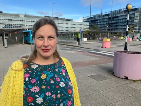 BBC/Amy Holmes Photo of a blonde woman with her back tied. She wears a yellow cardigan that covers a dark green dress with flowers of different colors and sizes.