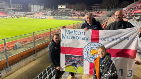 A boy and his family holding a Leicester City St George's Cross flag in the stands of a stadium