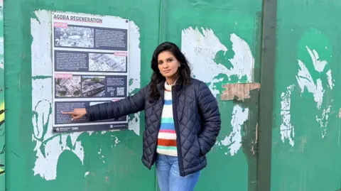 MK Conservatives Shazna Muzammil with long dark hair wearing a black jacket over a multi-coloured jumper, pointing at a notice on the side of green fencing which says "Agora regeneration".