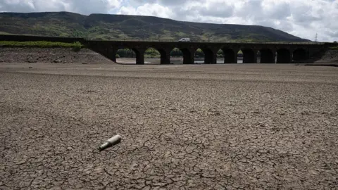 Parched, cracked land in the foreground. An empty bottle lies on the surface. In the background is a bridge with a white van driving across.