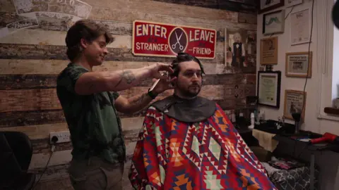 A barber with a green shirt and brown trousers stood and cutting the hair of a man who is wearing a patterned gown and sat in a chair having his hair cut in a barbershop.