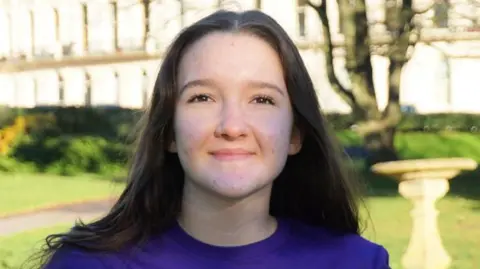 A teenage girl, wearing a dark purple t-shirt over a long sleeve black shirt. She is standing at a park.